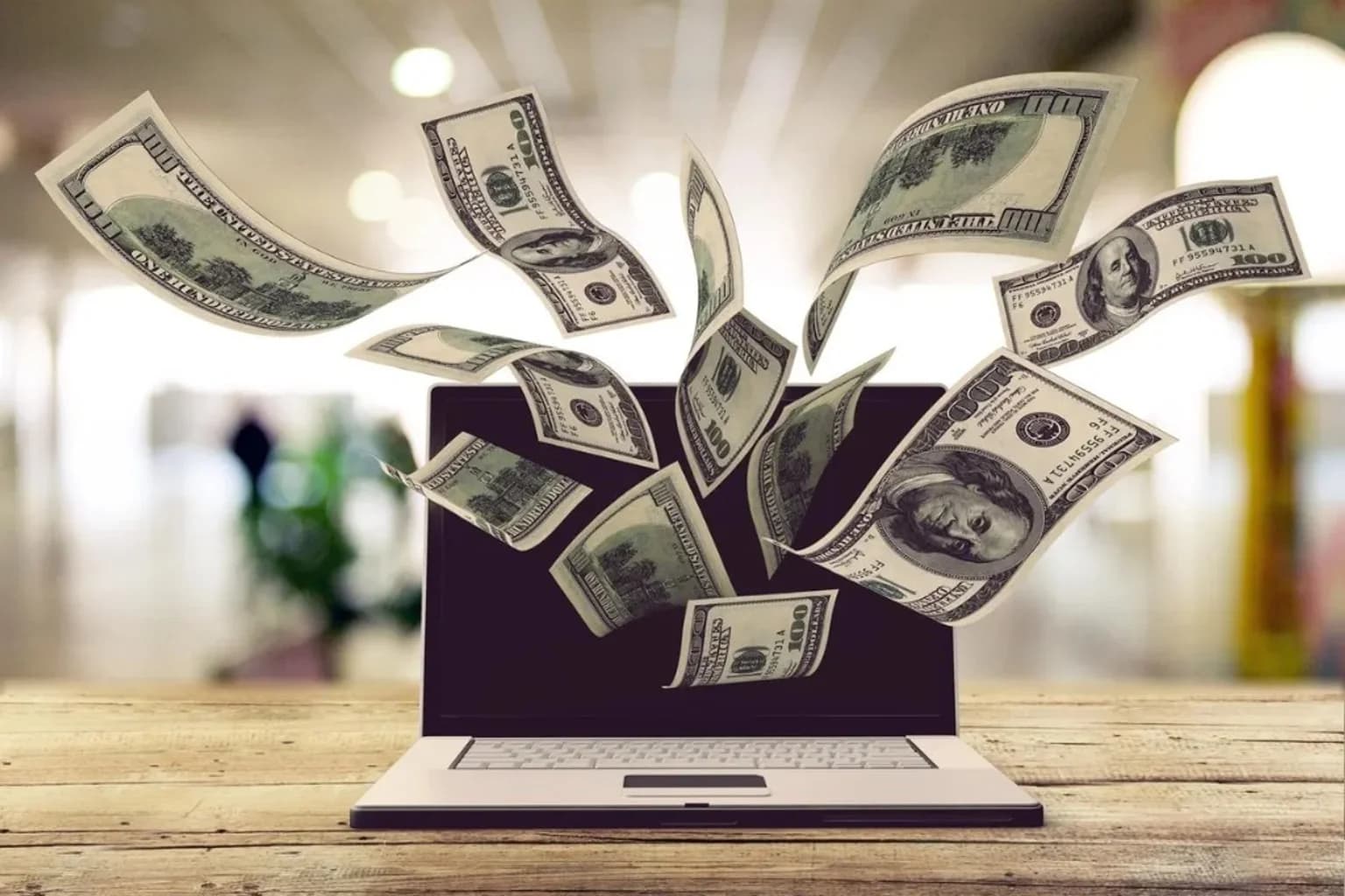 Laptop on a wooden desk with hundred-dollar bills rising from the screen